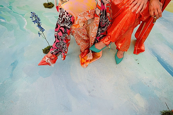 Wedding shoe detail with colorful wedding heels, pointed-toe pumps beside patterned outfits on a painted floor with moss patches and a blue flower stem