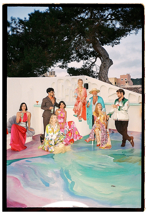 Wedding party portrait with a bridal party by pool, posing in colorful patterned outfits holding cocktails against a white stucco wall