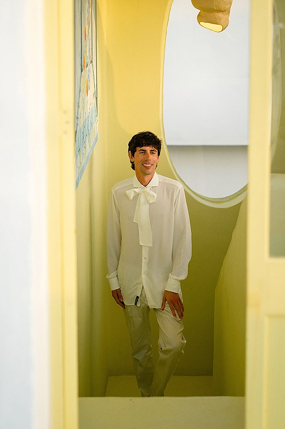 Groom portrait in an oversized bow tie, wearing a white dress shirt and light trousers in a yellow stairwell with pendant light