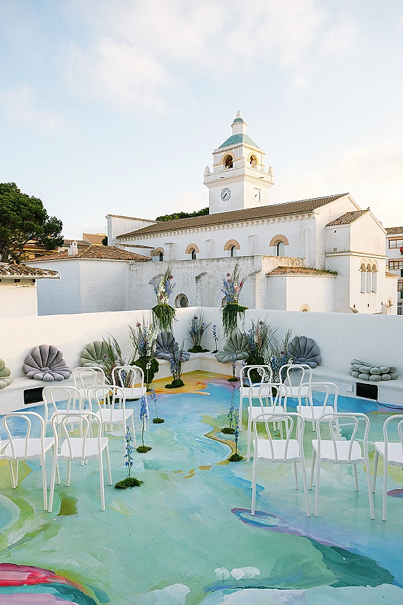 Ceremony setup with white ceremony chairs and blue florals lining the aisle on moss bases in a stucco courtyard under tiled rooftops