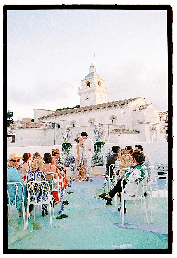 Ceremony moment at an outdoor wedding ceremony with couple at altar, bouquet and floral aisle arrangements on a rooftop terrace by a white church clock tower