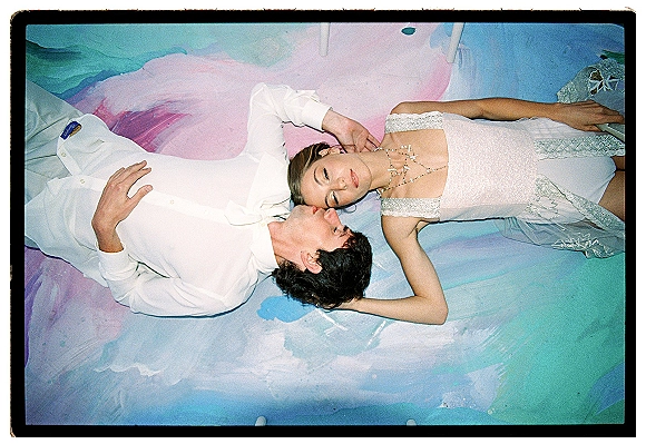 Couple portrait of bride and groom lying down in an overhead wedding portrait on a colorful painted floor with pastel backdrop and lace dress details