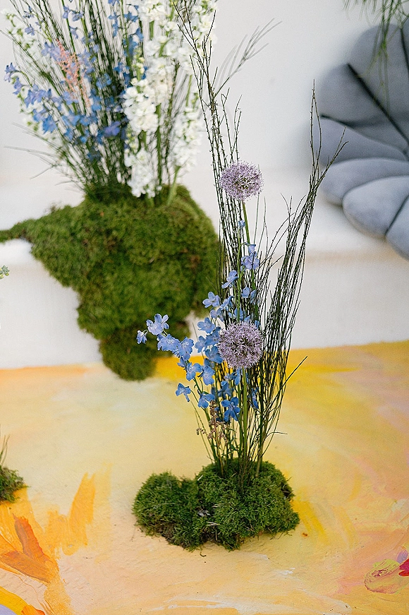 Floral centerpiece with moss centerpiece base, wildflowers, blue and white blooms, purple allium, and twigs on a painted surface by a neutral wall