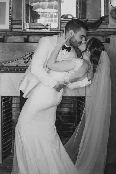 Wedding kiss portrait of bride and groom in a dramatic dip, her long veil and lace sleeves framed by an indoor fireplace mantel