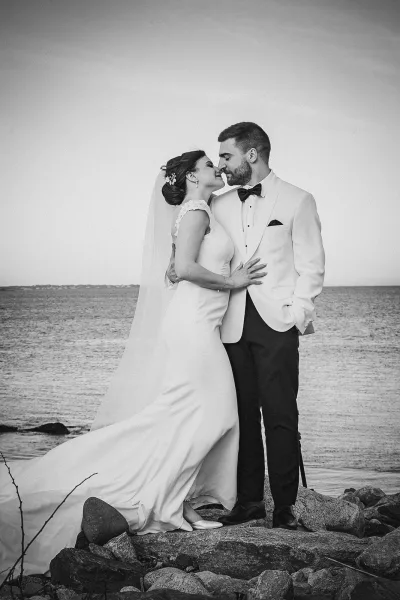 Wedding couple portrait in black and white, bride and groom by ocean touching foreheads on rocky shoreline, veil flowing in sea wind