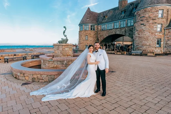 Couple portrait of bride and groom embracing, her long veil trailing beside his white tuxedo jacket, with castle courtyard and ocean view behind.