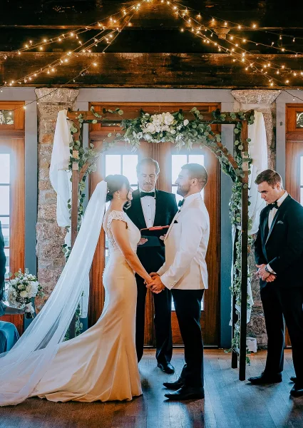 Wedding vows as bride and groom hold hands beneath a greenery wedding arch with string lights in a rustic stone-and-wood indoor venue