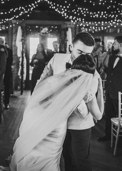 First dance as groom kisses bride’s forehead, her long veil and lace dress flowing under string lights in an indoor reception space