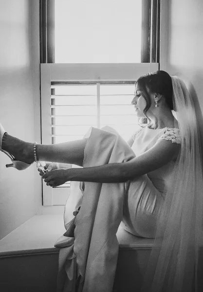 Bridal getting ready as bride putting on heels by a window seat, veil and lace-sleeve wedding dress lit by soft window light