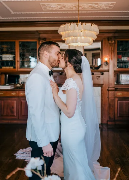 Wedding kiss as bride in lace dress and veil embraces groom in white tuxedo jacket, ring visible, in a wood-paneled bar interior