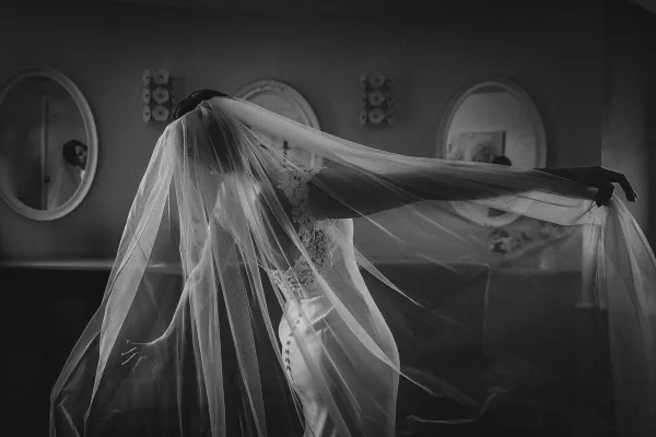 Bridal portrait of a bride with veil, arms outstretched in a lace wedding dress, reflected in wall mirrors under ceiling lights indoors