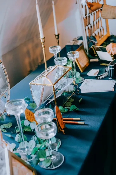 Wedding guest book on a dark teal table with eucalyptus garland, taper candles, glass card box, and champagne coupes by a wood railing