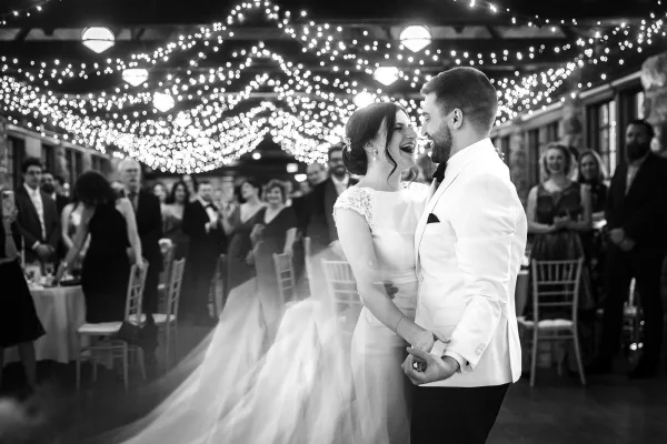 First dance as bride in lace cap sleeve wedding dress and groom in white dinner jacket twirl under string lights, guests watching indoors