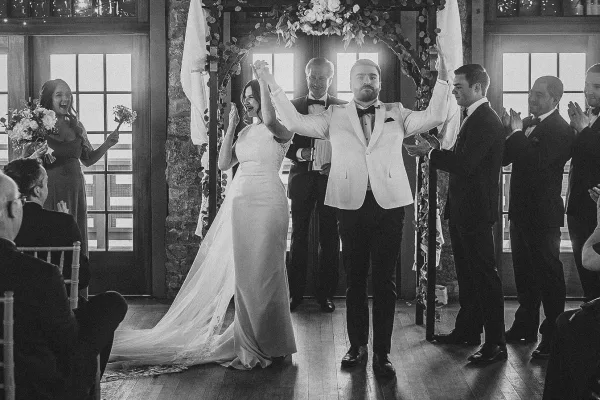Wedding recessional as bride and groom walk the aisle with raised hands under a floral arch, guests cheering in a bright indoor venue with windows