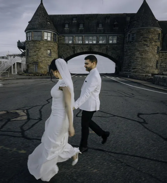 Couple portrait of bride and groom walking hand in hand, her long veil flowing, beside a stone castle archway under overcast sky