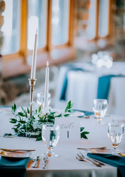 Reception tablescape with wedding table centerpiece of taper candles in brass candlesticks, greenery garland, and white flowers by large windows