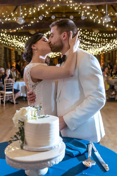 Wedding cake cutting as bride and groom lean in for a cake cutting kiss beside a white buttercream cake under reception string lights