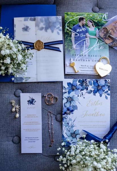 Invitation suite with blue floral wedding invitations, vellum wrap, wax seal, navy ribbon, rings and jewelry on gray fabric backdrop