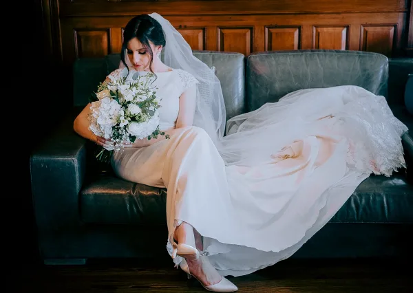 Bridal portrait of a bride sitting on couch, looking down with veil and white bouquet, lace sleeves and heels by wood-paneled wall