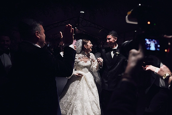 Wedding recessional as newlyweds walking aisle hand in hand, bride in lace dress and veil laughing beside tuxedoed groom in indoor corridor at night
