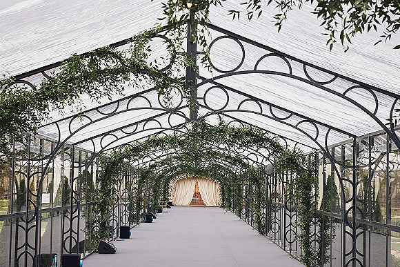 Ceremony aisle design with a greenery wedding aisle framed by metal arches, leading to a draped backdrop inside a glass pavilion tent