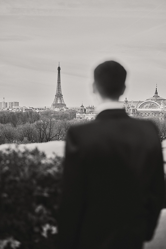 Groom portrait from behind in a tuxedo with bow tie, looking away on a rooftop terrace with city skyline and tower landmark under overcast sky
