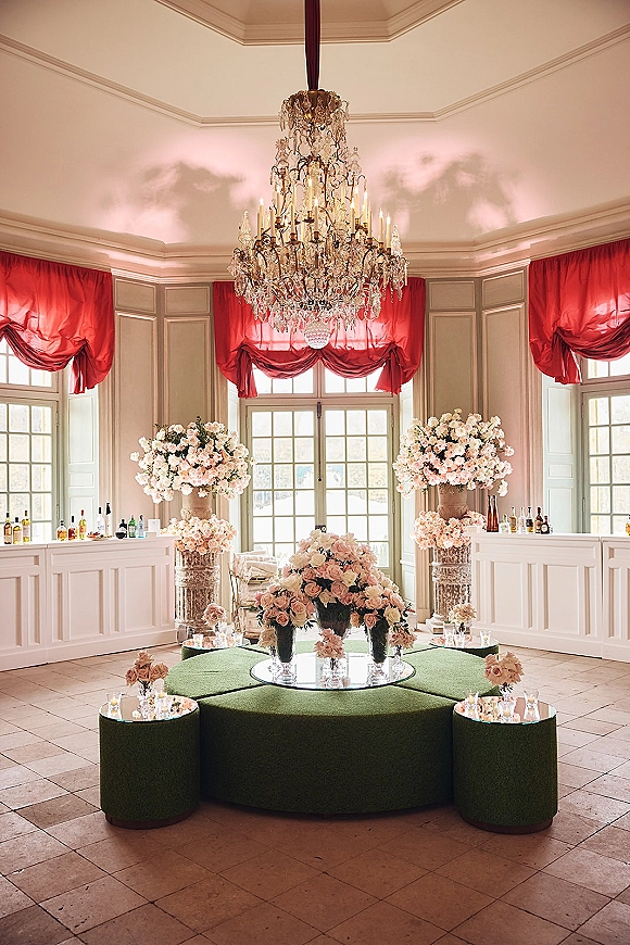 Wedding lounge decor with ottoman seating, mirrored coffee tables, rose florals, votive candles, and a crystal chandelier in a grand ballroom