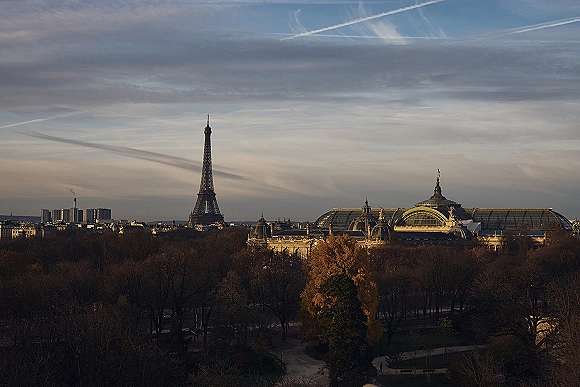 City skyline with Eiffel Tower view glowing in sunset light above treetops, a historic glass-roof building, and a cloudy sky