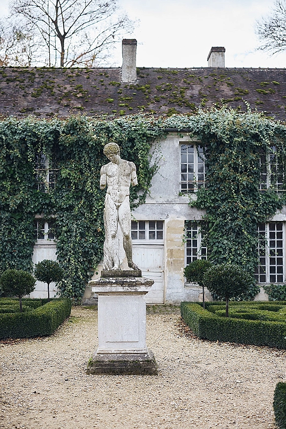 Garden statue on a stone statue pedestal centered along a gravel path, framed by boxwood hedges and an ivy-covered building backdrop