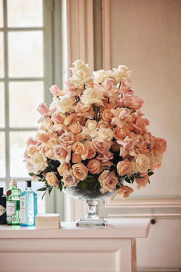 Rose centerpiece of pink and ivory roses with greenery in a glass pedestal bowl on a bar counter, with bottles and window panes behind