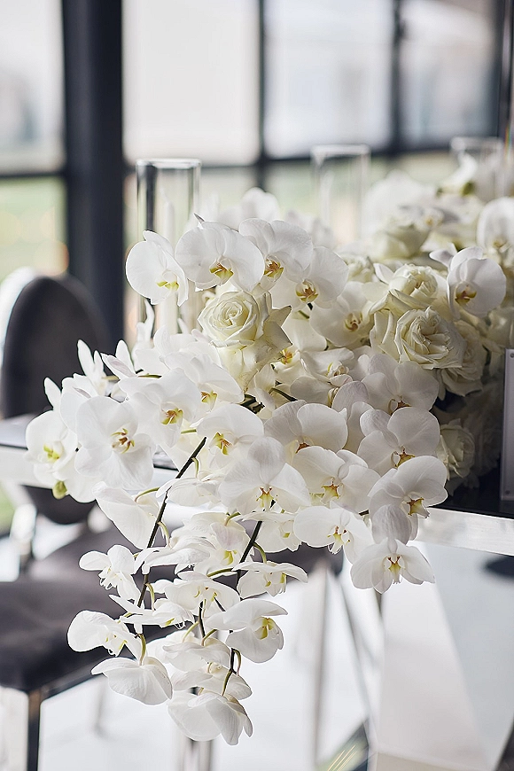 Wedding centerpiece with white orchid centerpiece blooms and white roses in clear cylinder vases, cascading softly in window light near blurred chairs