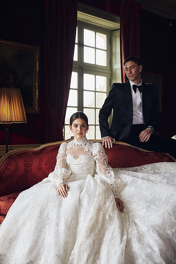 Couple portrait of bride in a high neck lace wedding dress seated on a sofa as the groom stands behind in a black tux by a large window