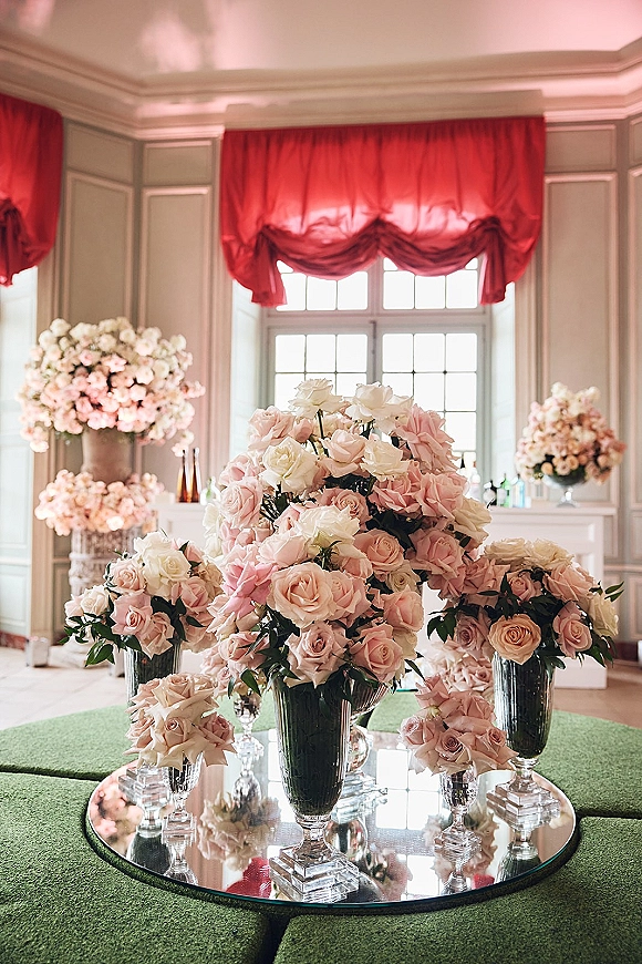 Wedding floral centerpieces of pink rose centerpieces in glass vases on a mirrored tray atop a cocktail table, set in a paneled ballroom lounge