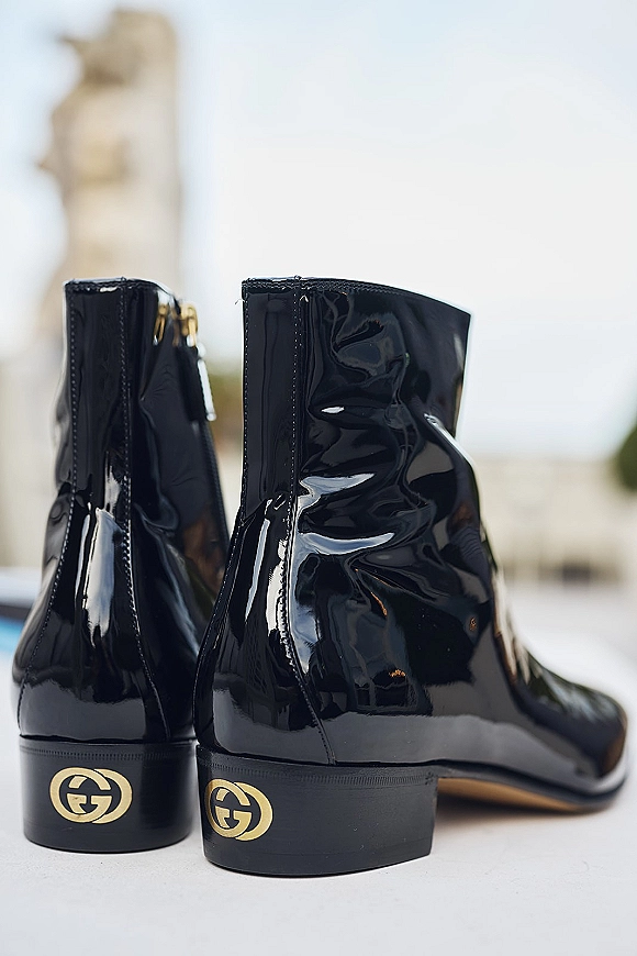 Groom wedding boots in shiny black patent leather with a gold logo detail, photographed against blurred outdoor architecture and sky