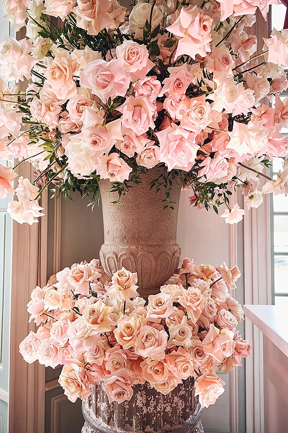 Rose floral arrangement with blush and ivory roses and greenery in a stone urn on a distressed metal planter by indoor windows