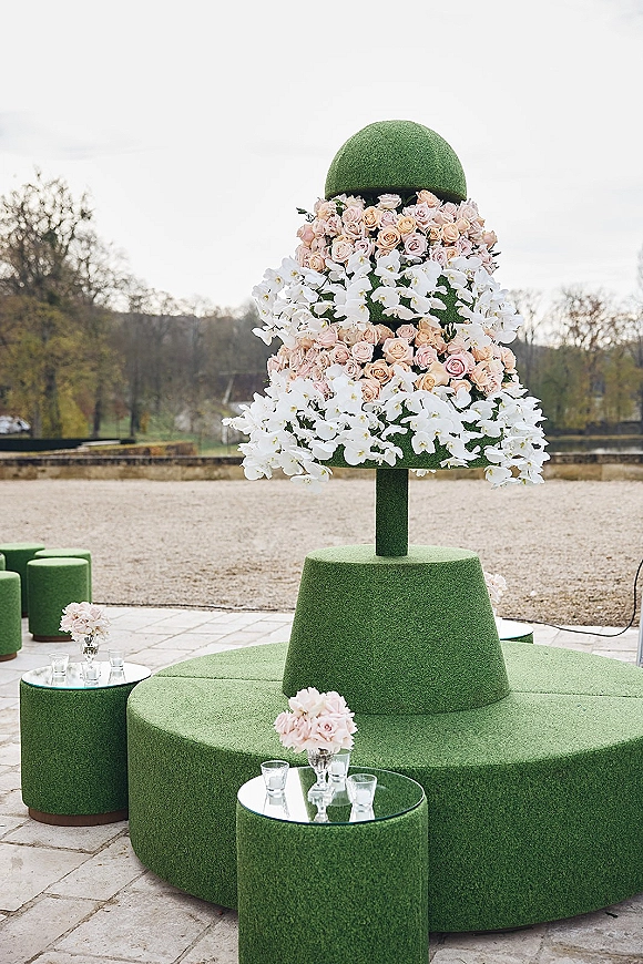 Wedding floral installation with a ceremony floral pillar of pink roses, white orchids and greenery beside green ottomans on a stone patio