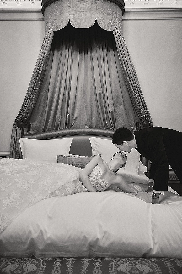 Wedding couple portrait of bride and groom on bed as he leans in for a kiss, strapless lace dress and tuxedo in hotel bedroom