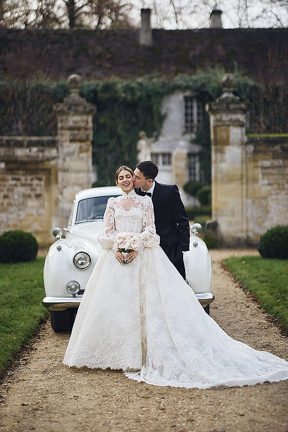 Couple portrait of bride in long-sleeve lace wedding dress kissing groom by a vintage wedding car at stone gate estate driveway with ivy wall