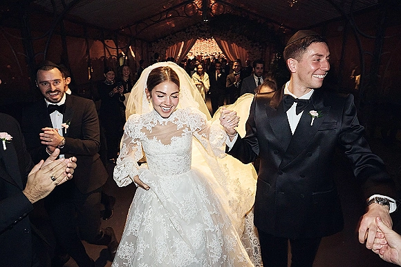 Wedding recessional as newlyweds walk down the aisle holding hands, bride in lace dress and veil, guests cheering in a warm-lit tent