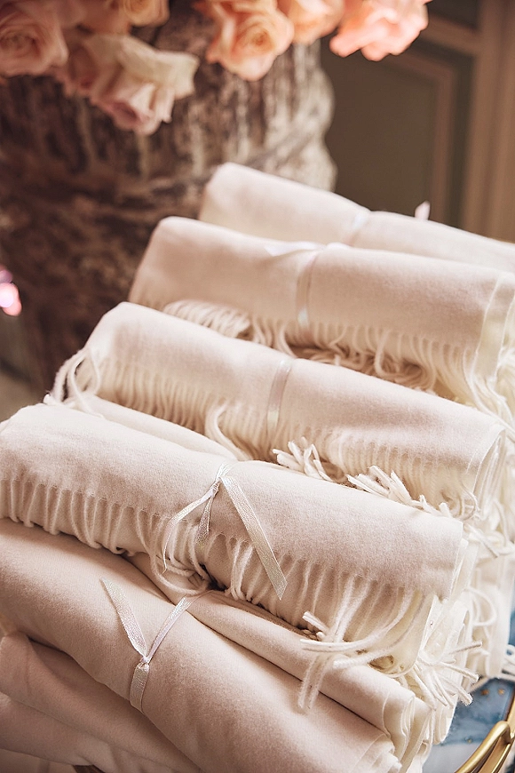 Wedding reception blankets rolled and tied with ribbon, fringe tassels visible on a tabletop comfort station with blush rose accents