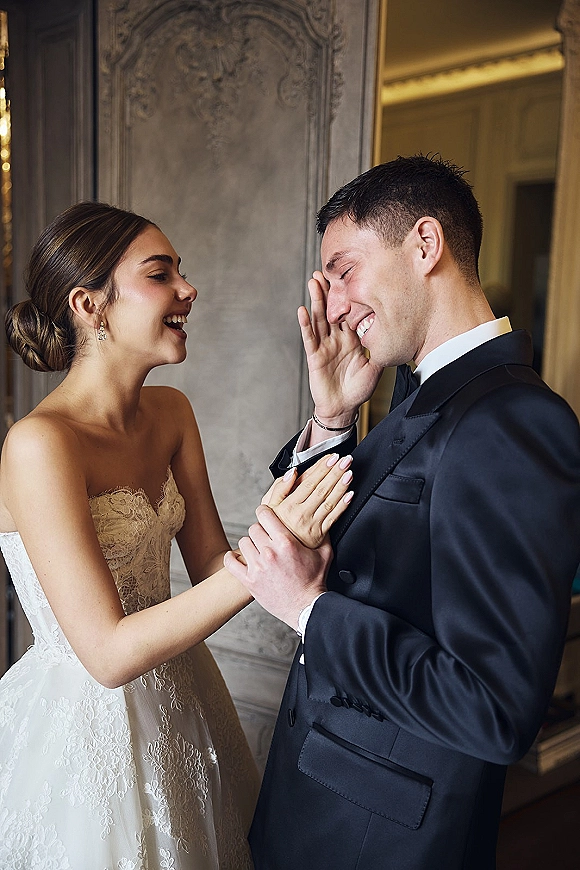 Couple portrait of bride and groom laughing while holding hands, her strapless lace wedding dress glowing by ornate wall paneling indoors