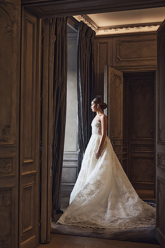 Bridal portrait of a bride in a strapless lace wedding dress with a ball gown skirt, posing by a tall window in an ornate room