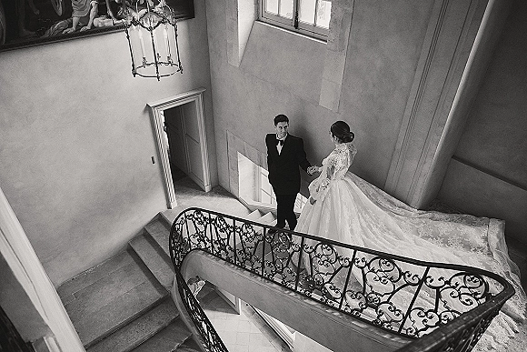 Couple portrait of bride and groom staircase pose, holding hands as her long train falls on stone steps under chandelier light