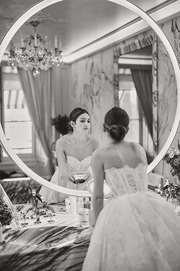 Bride getting ready at a bridal vanity mirror, wearing a strapless lace wedding dress with low bun, reflected in a marble bathroom.