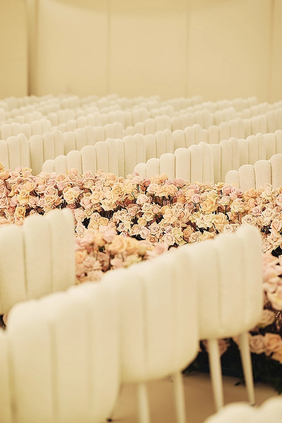 Ceremony aisle decor with a floral aisle runner of blush roses and rose petals between white chairs in an indoor space with cream walls