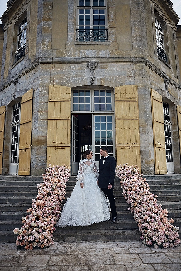 Couple portrait of bride and groom on steps, gazing at each other in lace-sleeved dress and tuxedo, framed by pink roses at a historic facade