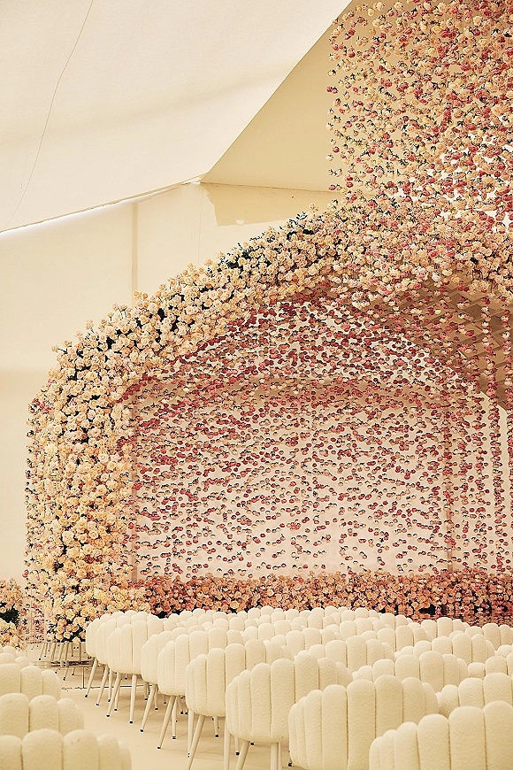 Ceremony setup with wedding ceremony seating beneath a hanging flower installation, white chairs facing a rose floral arch inside a tent