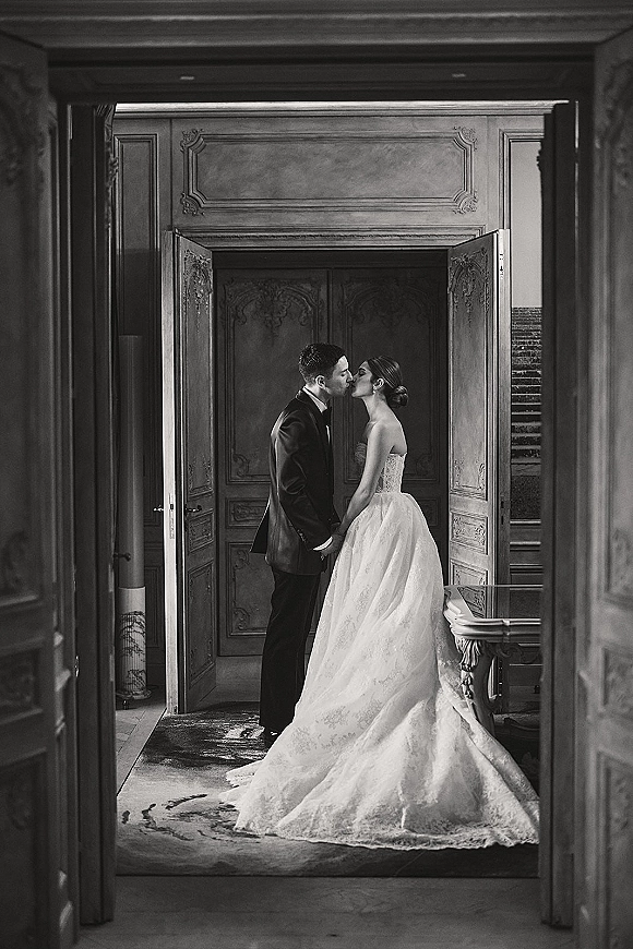 Wedding kiss portrait of bride and groom kissing in a doorway, her strapless lace gown and train beside his tuxedo, ornate doors behind