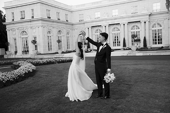 Couple portrait of bride twirling in a strapless wedding dress and veil as groom in tuxedo holds bouquet on a formal garden lawn