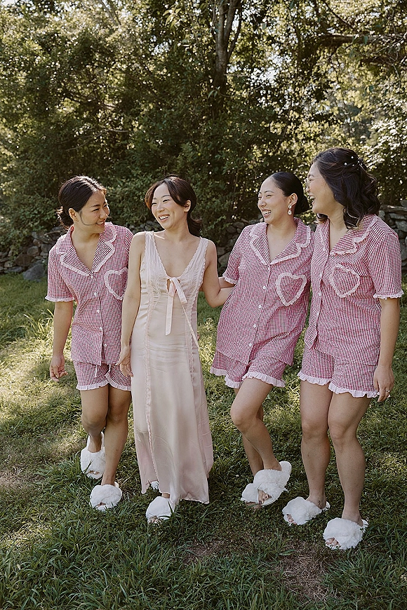 Bridesmaids getting ready in matching bridesmaid pajamas, walking together on a sunny lawn with trees and a stone wall behind them